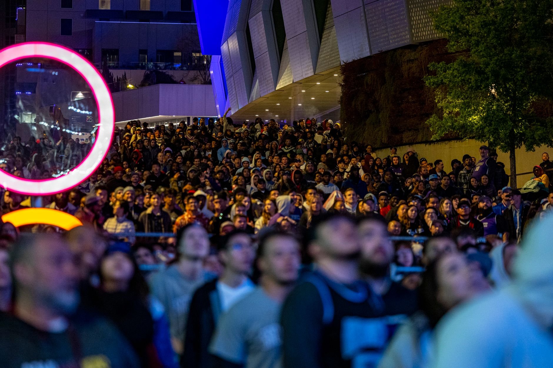 A lively scene of a large crowd gathered at an outdoor event at night, illuminated by vibrant neon lights.